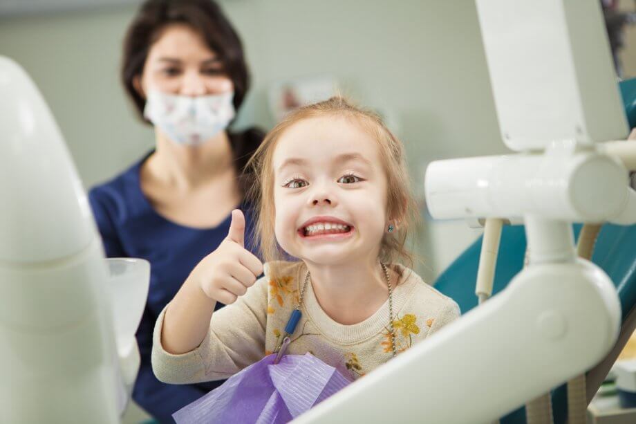 Photograph of a young girl with straight red hair smiling and giving a thumbs up while at the dentist.