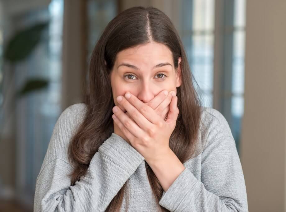 Woman wearing a gray long-sleeved shirt with her hands over her mouth.